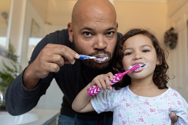A father and daughter brush teeth in the same mirror.