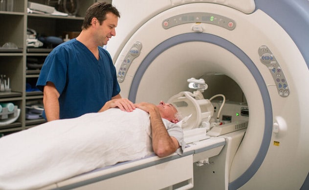 A health care provider stands by an MRI machine as a person lays on a table outside the machine.