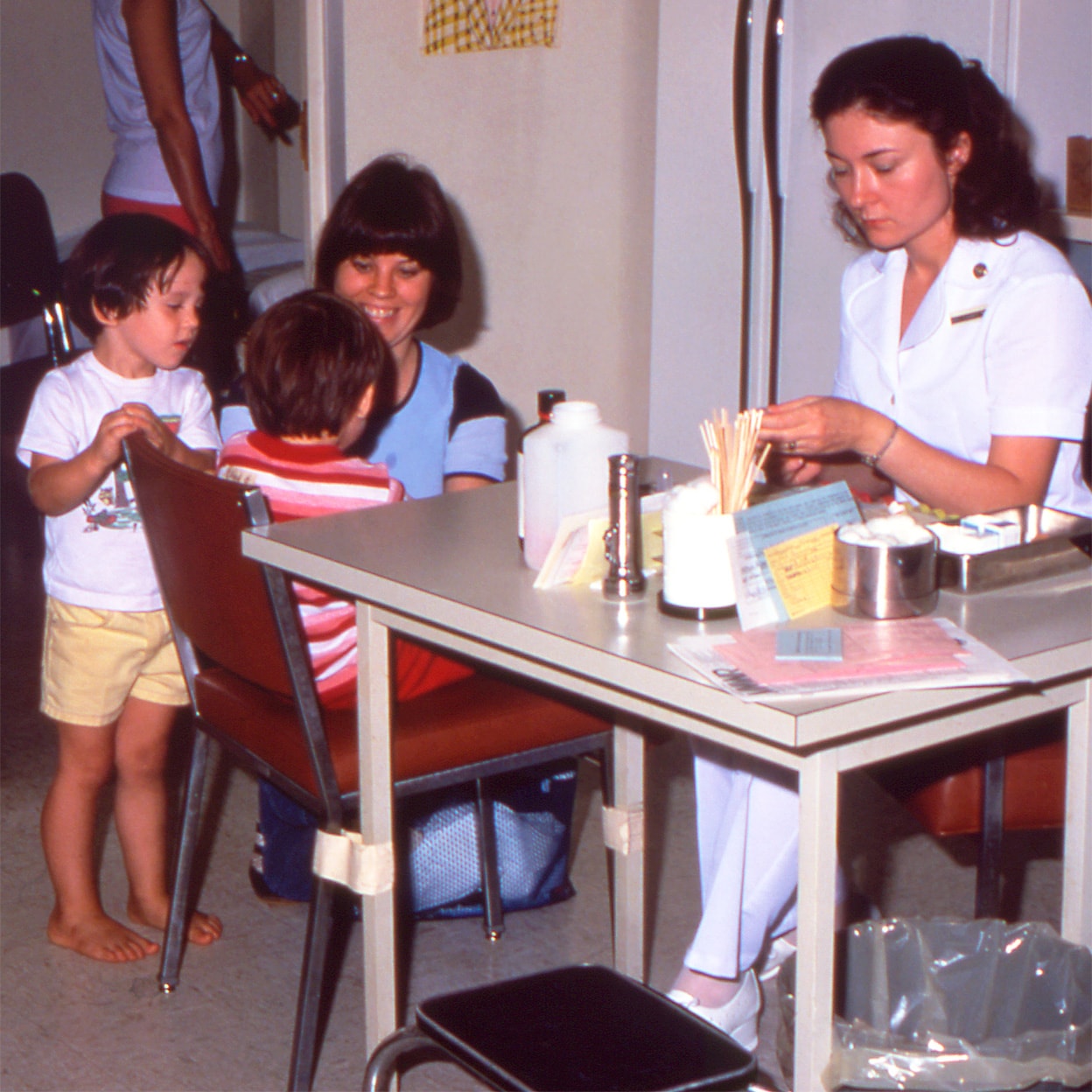A mother waits with two children as another child prepares to get a vaccine from a nurse.