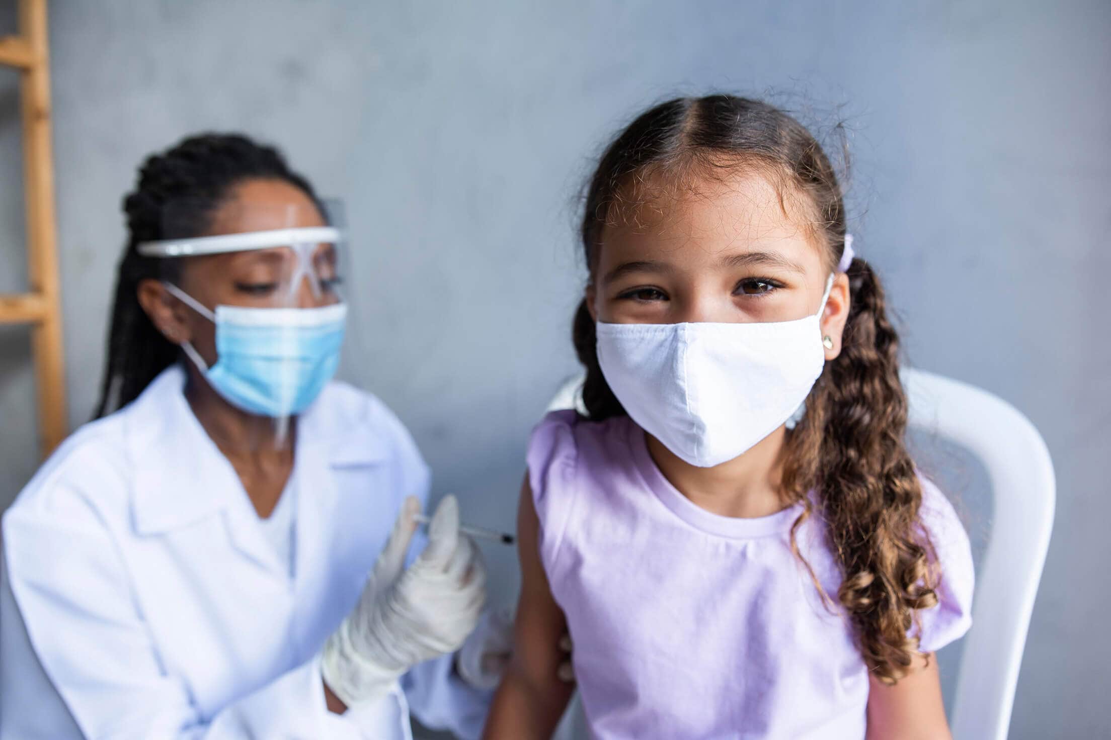 A young girl wearing a mask gets a COVID-19 vaccine from a health care professional wearing a mask and face shield.