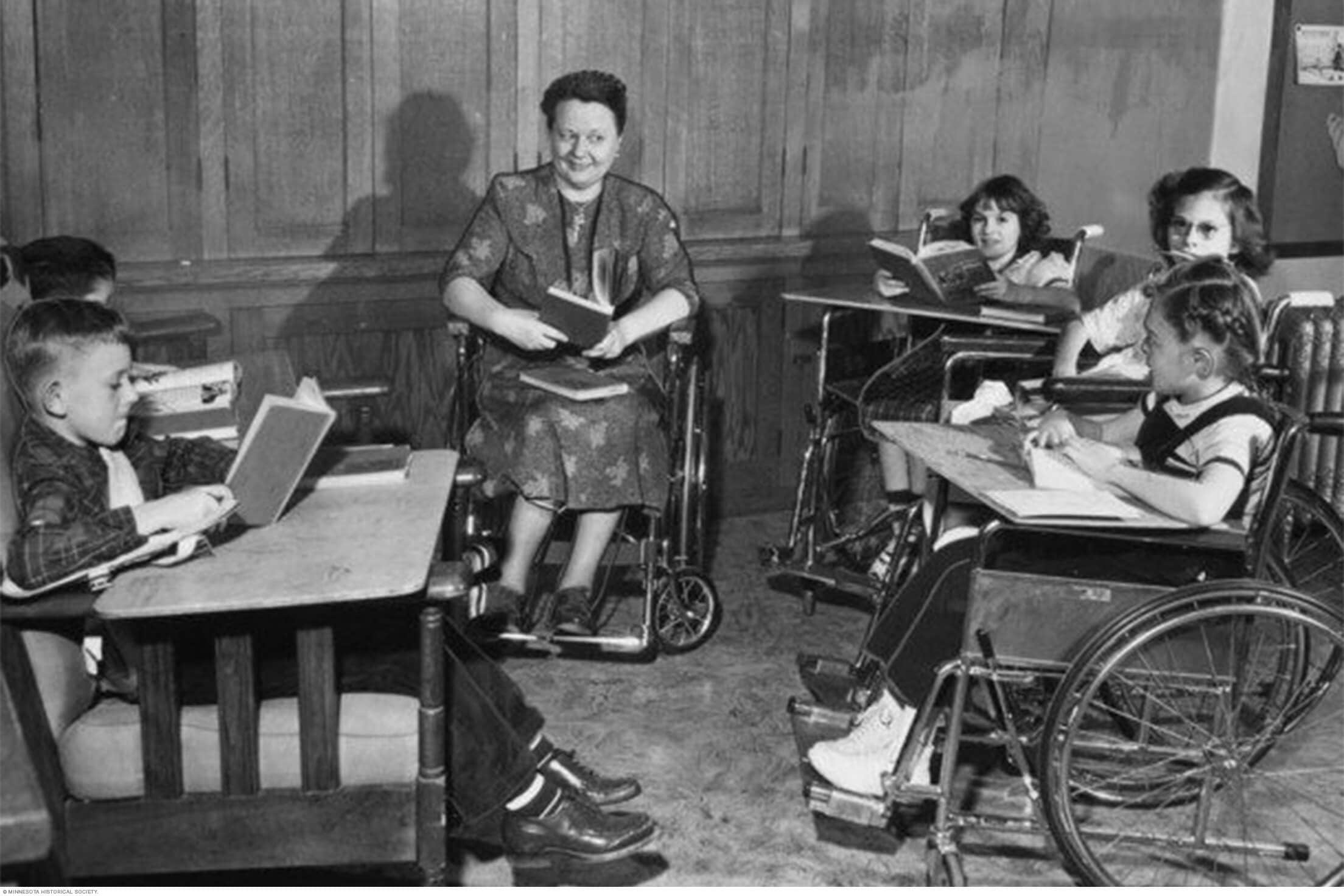 أطفال ومعلمة يجلسون في مقاعد متحركة ممسكين كتبًا ودفاتر في فصل دراسي في مستشفى سانت ماري
