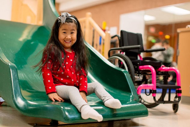 A young girl in the therapy center smiles at the bottom of a slide.