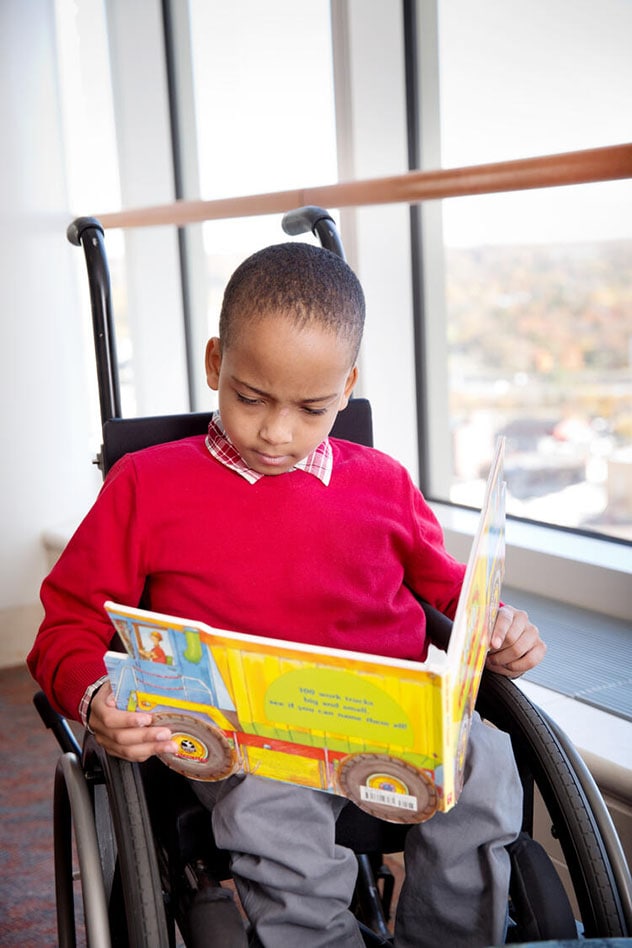A child in a wheelchair reads a book next to a sunny window.