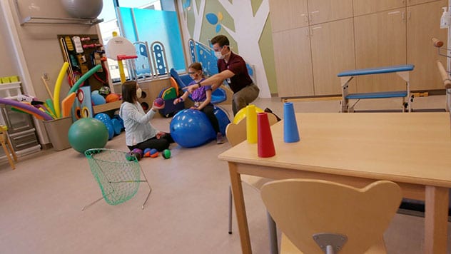 معالج متخصص في تأهيل الأطفال يجري جلسة علاج بالتمارين الرياضية مع طفلة ووالدتها في صالة الألعاب الرياضية العلاجية.