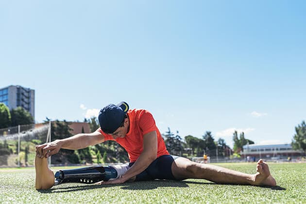 A young athlete pulls his prosthetic lower leg to stretch out on a field.