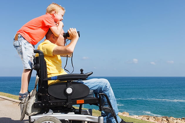 A therapy participant seated in an electric assisted chair and a smiling child companion look out to sea.