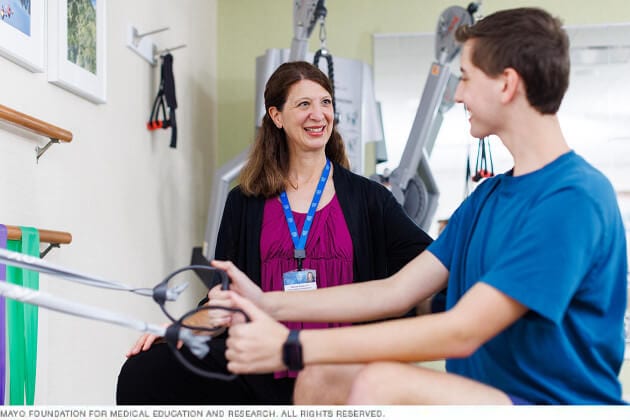 A young man and his therapist smile at one another as he exercises with elastic bands.