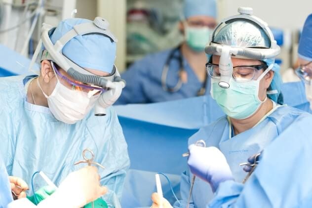 Two surgeons perform a procedure in an operating room with other team members in the background.