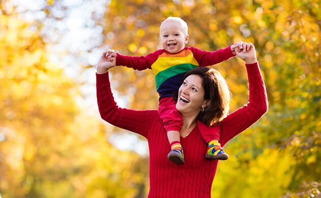 A woman has a smiling toddler up on her shoulders and she holds their hands for balance.