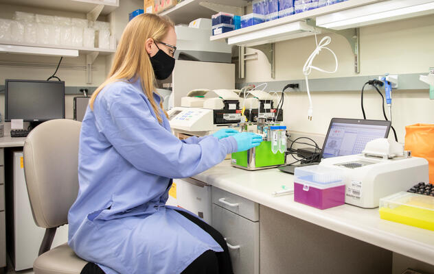 Photograph showing a masked and gowned lab technician sitting at a desk working with liquid samples